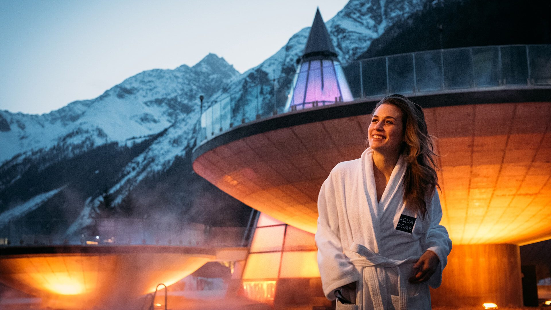 Eine Frau im weißen Bademantel lächelt in der Abenddämmerung im Freien. Hinter ihr sind schneebedeckte Berge und ein modernes, beleuchtetes Gebäude zu sehen, aus dem Dampf aufsteigt.