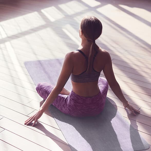 Eine Frau sitzt mit abgewandtem Gesicht auf einer Yogamatte in einem sonnendurchfluteten Raum und trägt einen grauen Sport-BH und violette Leggings.