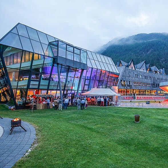 Modernes Glasgebäude mit bunten Lichtern neben einem traditionellen Gebäude, umgeben von Gras und Bergen. Menschen versammeln sich in der Nähe eines Zeltes auf dem Rasen.