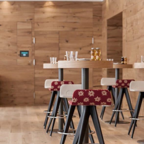 Barbereich mit hohen Holztischen und Hockern sowie rot gemusterten Kissen. Wände und Boden sind aus Holz und schaffen eine warme Atmosphäre.