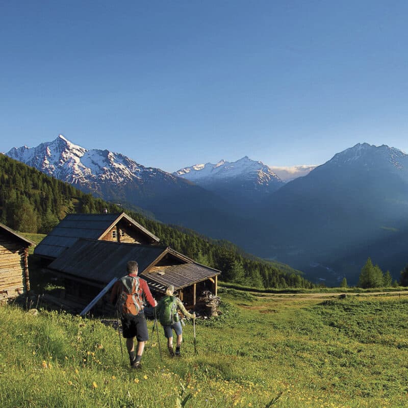 Zwei Wanderer mit Rucksäcken gehen auf einem Graspfad auf Holzhütten zu, umgeben von Bergen unter einem klaren blauen Himmel.