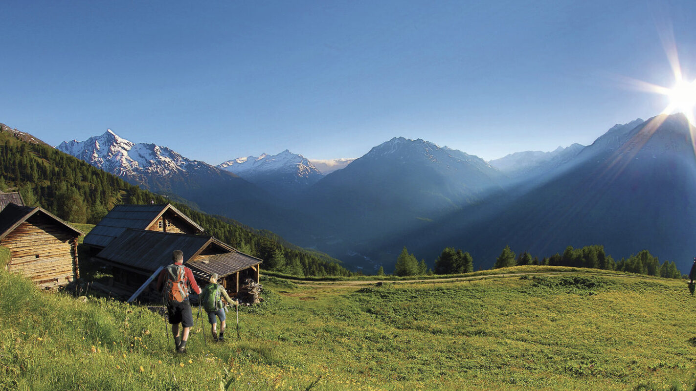 Two people stroll towards wooden cabins in a grassy summer meadow, framed by snow-capped mountains and a sunrise in the background.