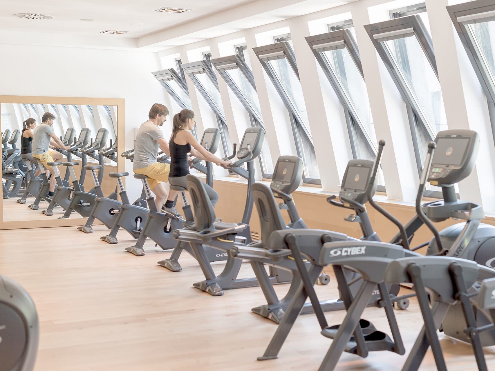 Zwei Personen trainieren auf Heimtrainern in einem hellen Fitnessstudio mit großen Fenstern und Spiegeln. Mehrere Trainingsgeräte stehen in einer Reihe.