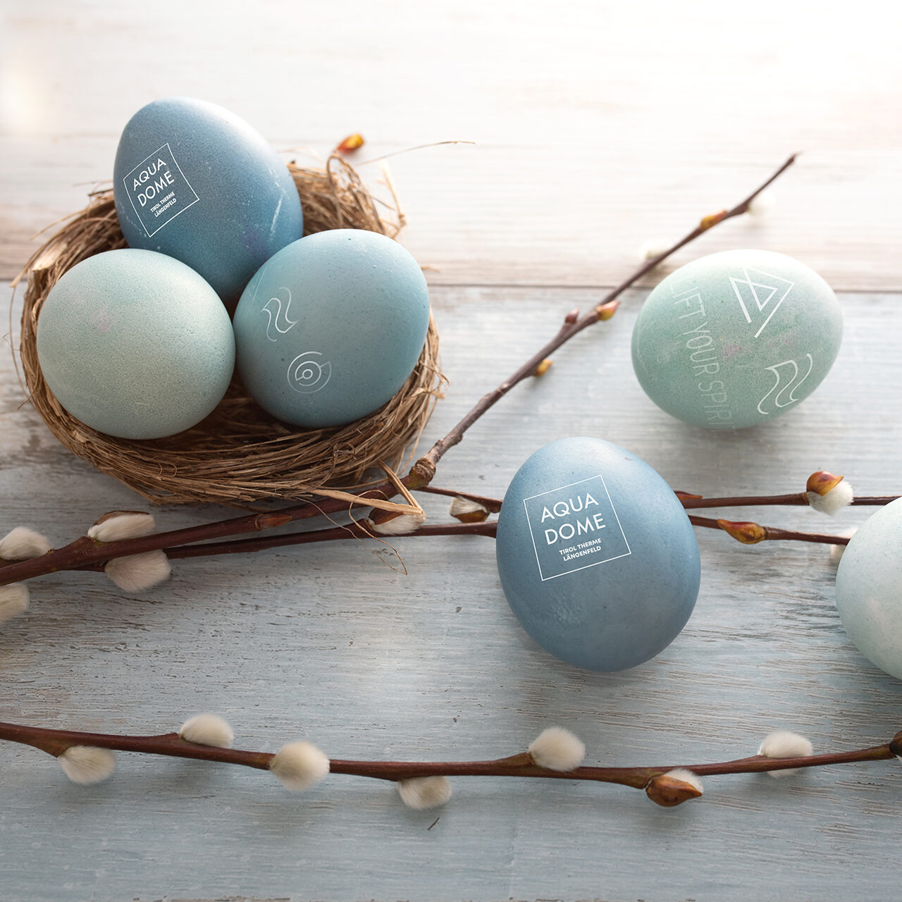 Vier blaue und grüne Eier in einem Nest und in der Nähe, mit geometrischen Mustern und Text, werden auf einer hellen Holzoberfläche mit Weidenkätzchenzweigen präsentiert.