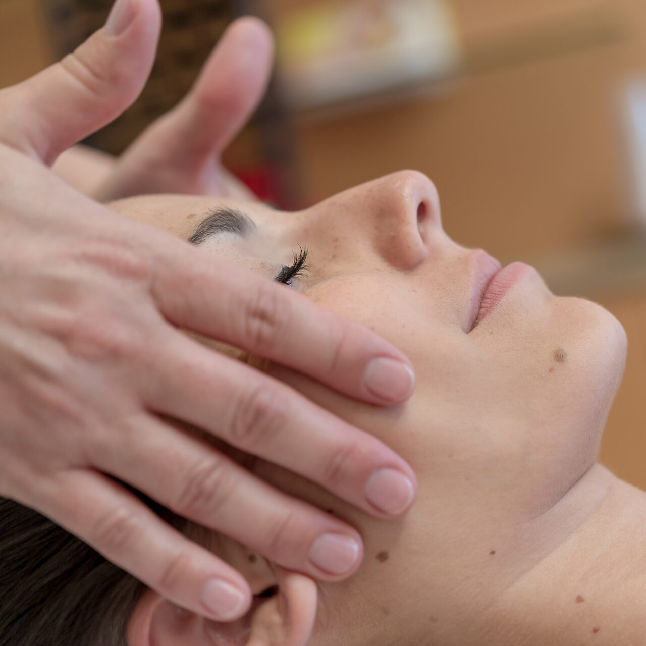 Nahaufnahme einer Person, die eine Gesichtsmassage erhält, wobei die Hände sanft auf ihre Schläfen und Wangen drücken und die Augen geschlossen sind.