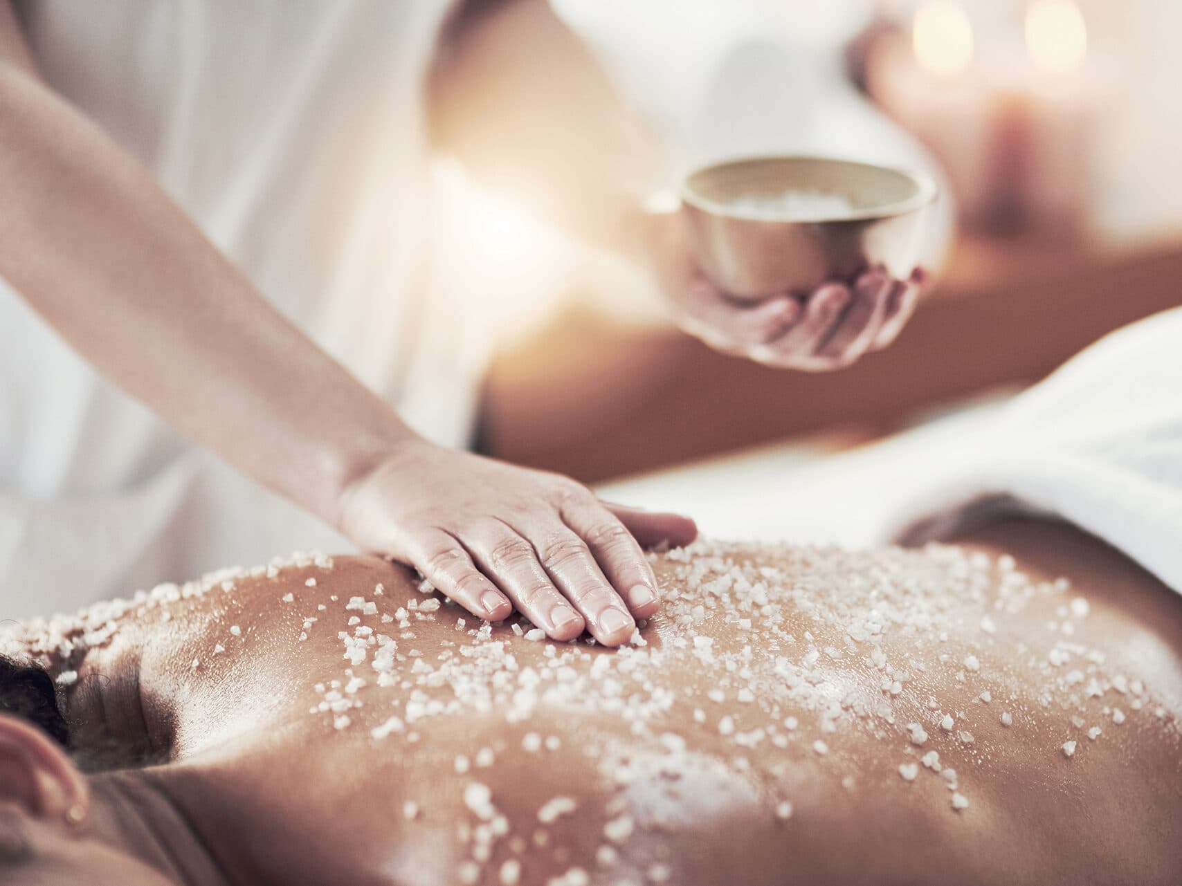 Eine Person erhält eine Salzpeeling-Massage. Die Hand des Therapeuten streicht Salz auf ihren Rücken. In der anderen Hand hält sie eine Schale mit Salz. Im Hintergrund sind Kerzen zu sehen.