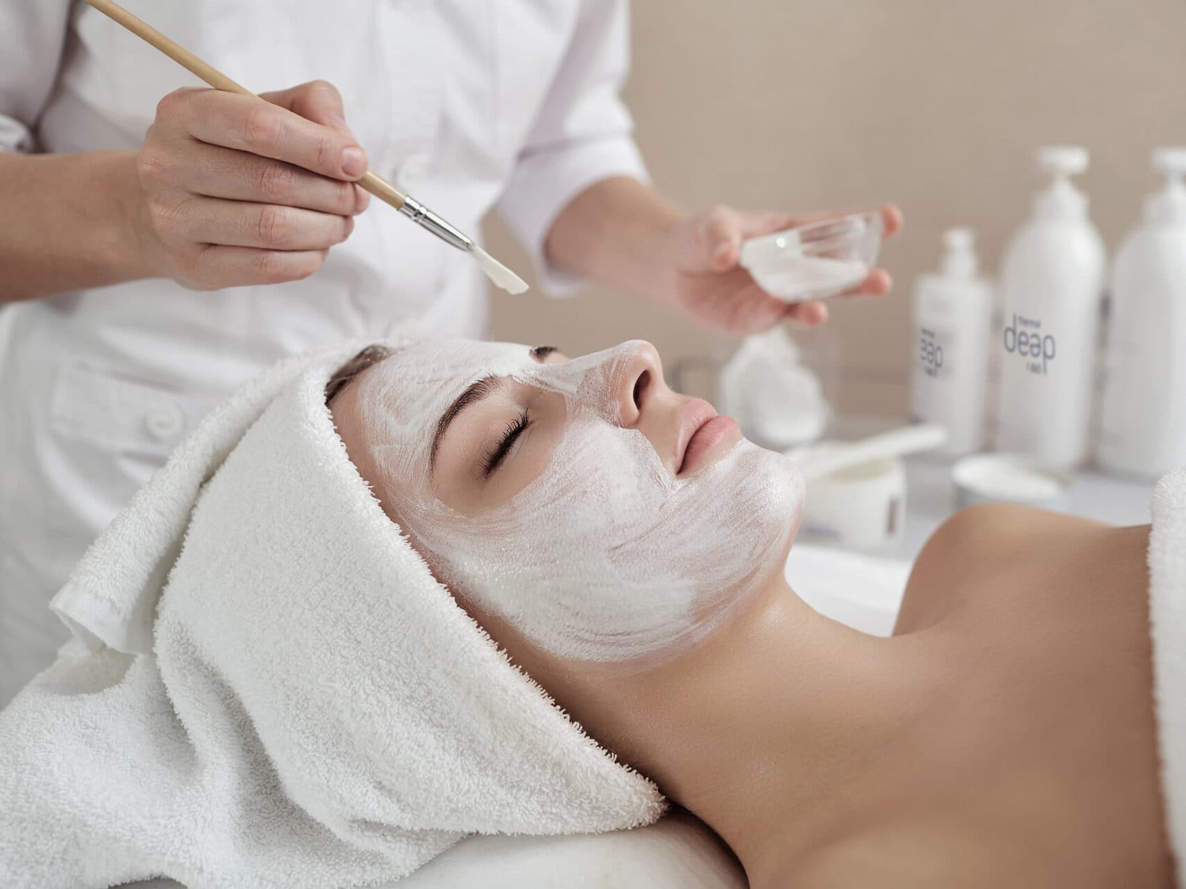 Person mit einem Handtuch auf dem Kopf erhält eine Gesichtsbehandlung. In einem Spa wird eine weiße Creme mit einem Pinsel aufgetragen.