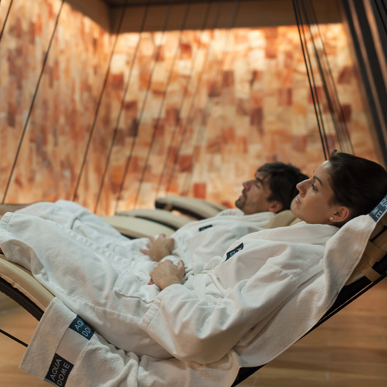 Zwei Menschen entspannen in weißen Bademänteln in Liegestühlen in einem Spa-Raum mit Holzböden und salzbedeckten Wänden.