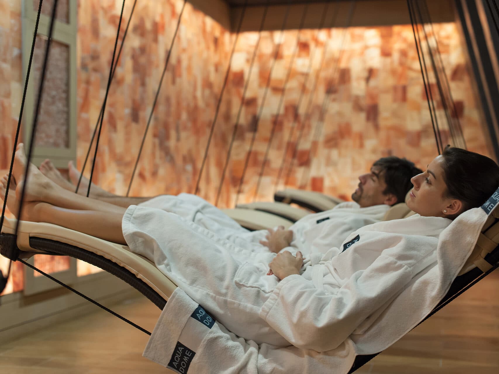 Menschen in weißen Bademänteln entspannen sich auf Hängeliegen in einem Spa-Raum mit salzbedeckten Wänden.