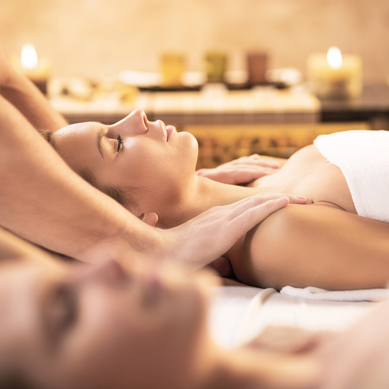 Zwei Personen erhalten eine Massage in einem Spa mit brennenden Kerzen im Hintergrund.