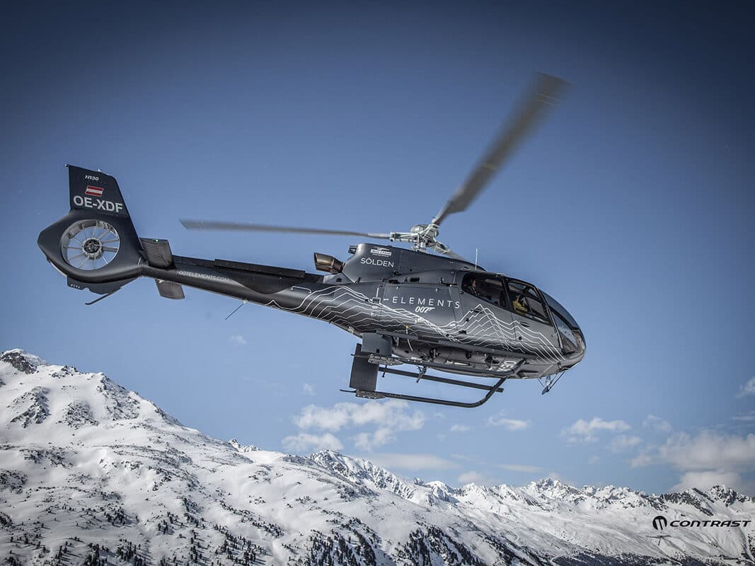 Ein Helikopter mit grafischem Design fliegt unter einem klaren blauen Himmel über eine verschneite Berglandschaft.