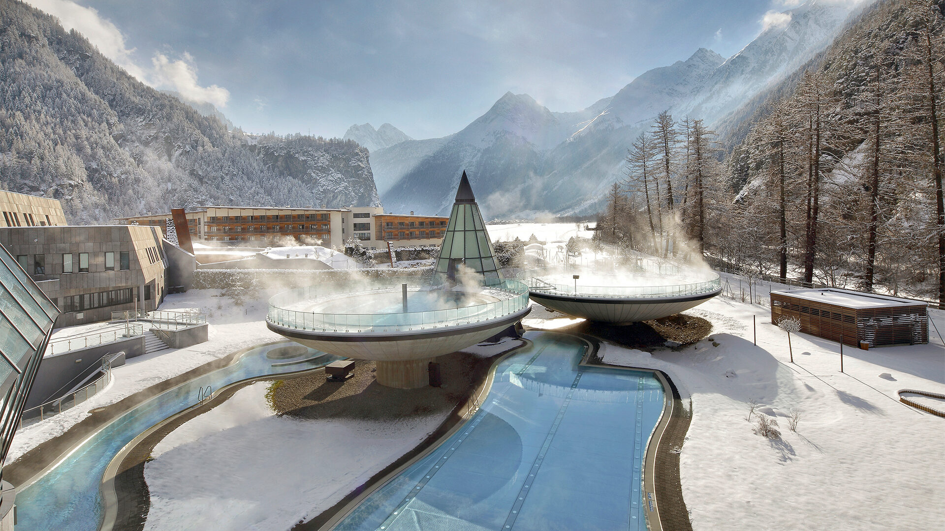 Inmitten einer verschneiten Berglandschaft bietet ein modernes Spa einen Anblick der Ruhe. Zwei runde, dampfende Strukturen sind von Wasserkanälen und Gebäuden umgeben, eingerahmt von Kiefern und klarem Himmel im Hintergrund.