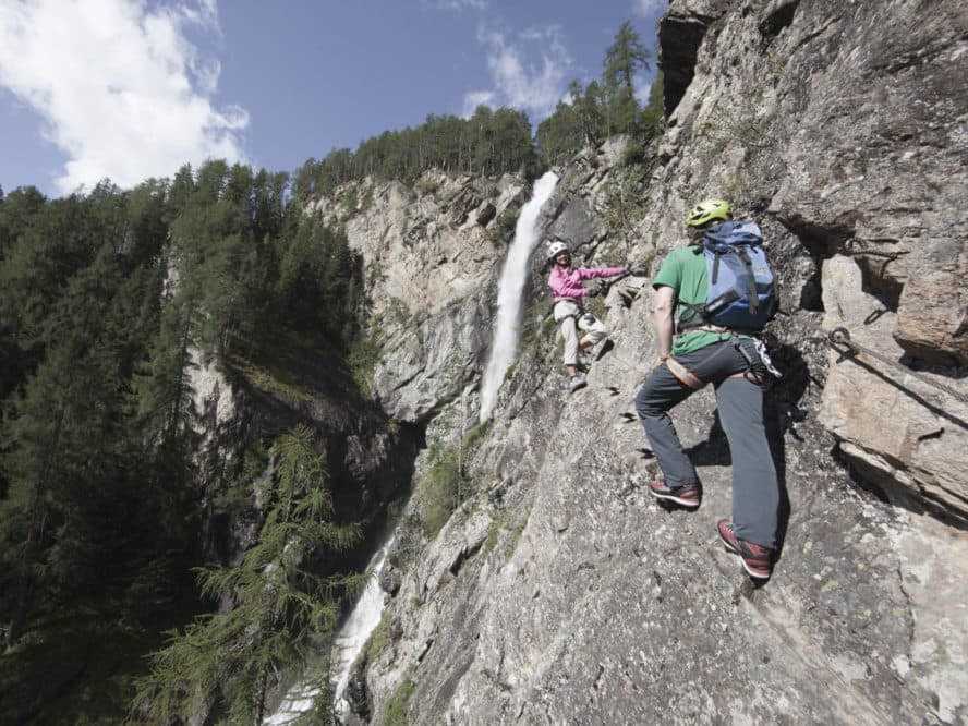Two climbers in helmets and gear navigate a steep rock face with a waterfall and forested landscape in the background under a clear sky.