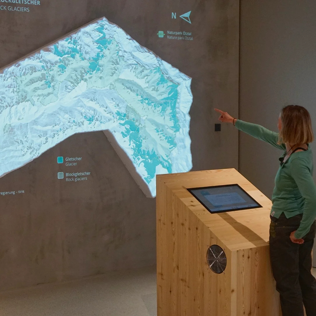 La persona indica una mappa topografica su una parete, con un podio di legno e un touchscreen in primo piano.
