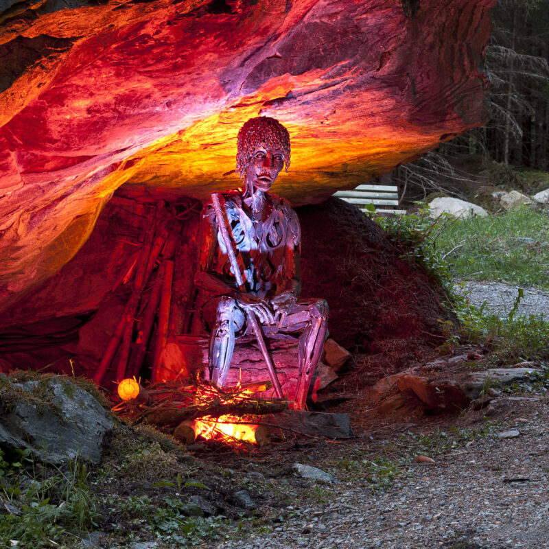 Metallskulptur einer Person, die in einer Felsenhöhle sitzt und von roten und orangefarbenen Lichtern beleuchtet wird.