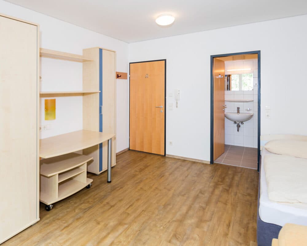 Minimalistisches Zimmer mit hellem Holzboden, Bett, Schreibtisch und Kleiderschrank. Eine offene Tür führt in ein kleines Badezimmer mit Waschbecken, Spiegel und gefliestem Boden. Die Wände sind weiß und mit Deckenbeleuchtung ausgestattet.