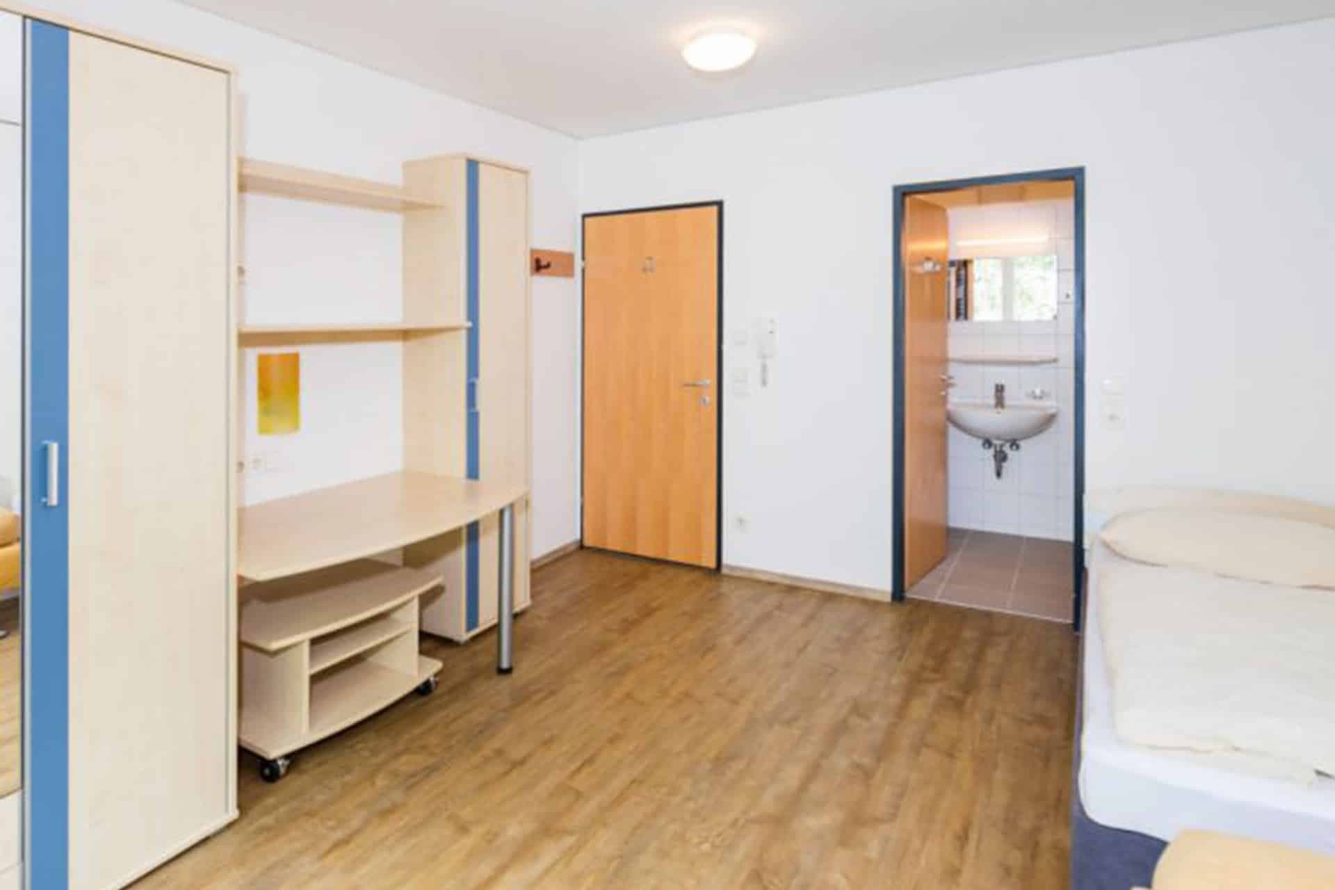 Ein schlichter, kleiner Raum mit Bett, Schreibtisch, Regalen und Bad. Helle Holzmöbel, eine geschlossene Tür, strahlend weiße Wände und Holzboden sind zu sehen.