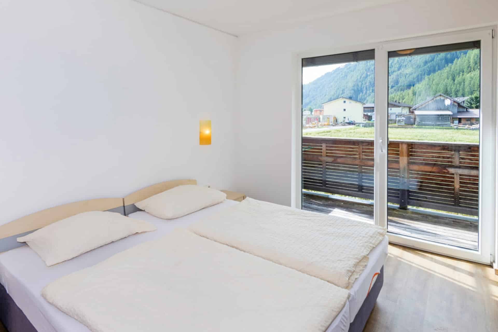 Ein minimalistisches Schlafzimmer mit einem Doppelbett, weißer Bettwäsche und einer teilweise geöffneten Schiebetür, die auf einen Holzbalkon mit malerischer Aussicht auf die Berge führt.