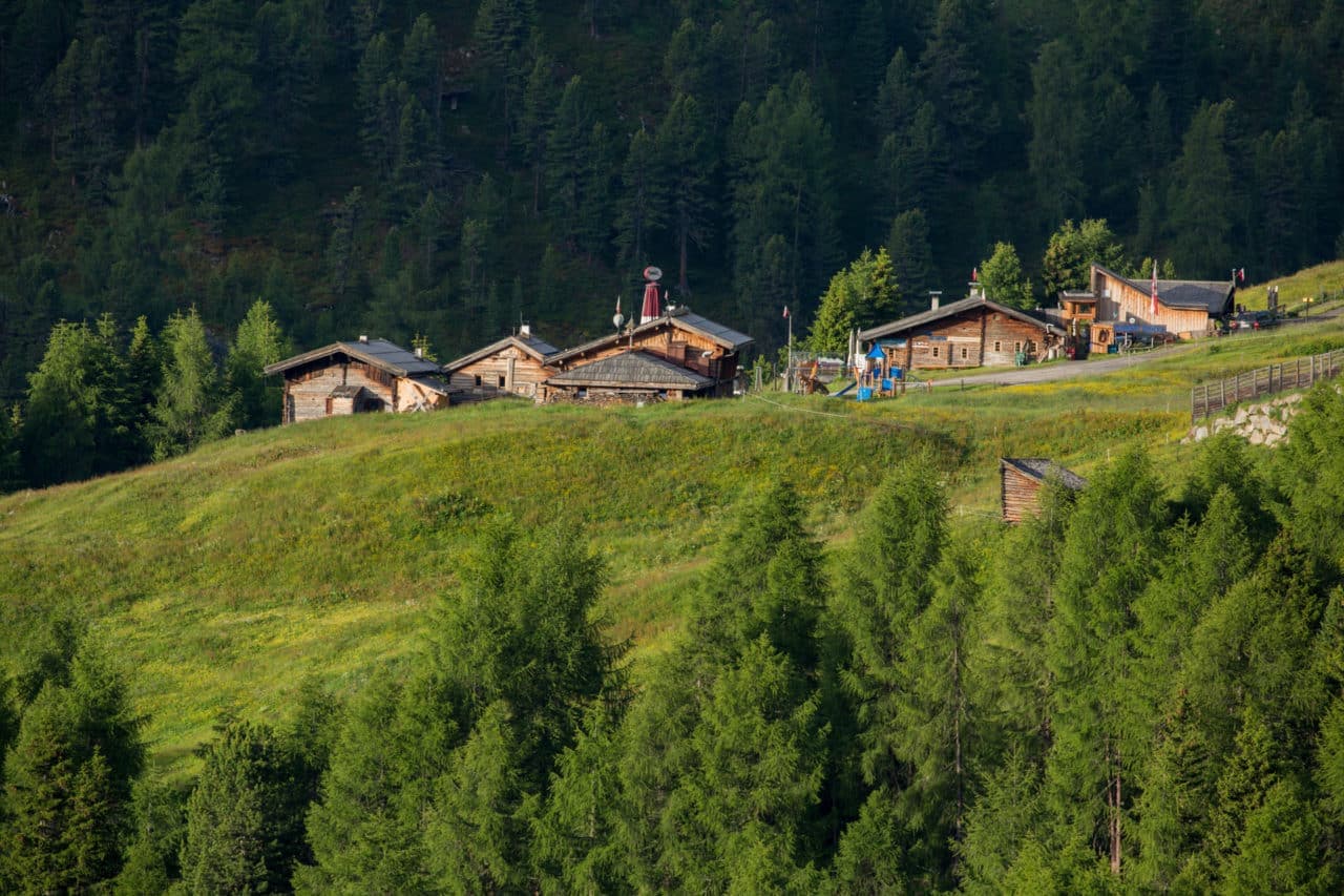 Die Holzhütten liegen verstreut auf einem grasbewachsenen Hügel, umgeben von dichtem Wald.