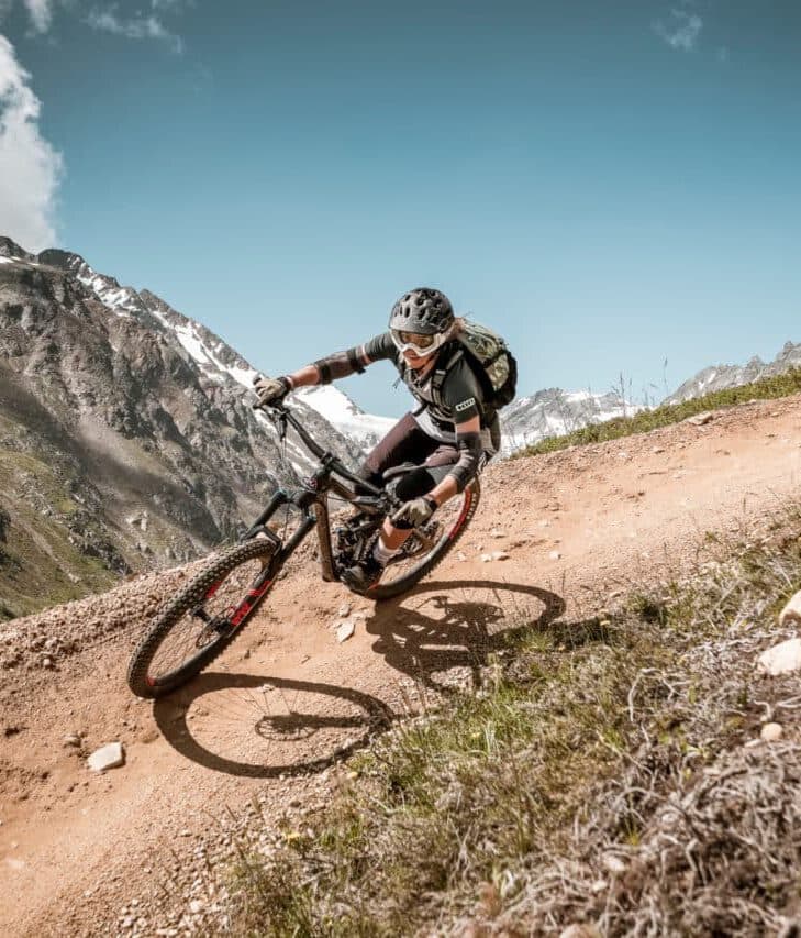 Eine Person fährt an einem sonnigen Tag mit dem Mountainbike einen Feldweg hinunter, umgeben von schroffen Bergen und Gras.