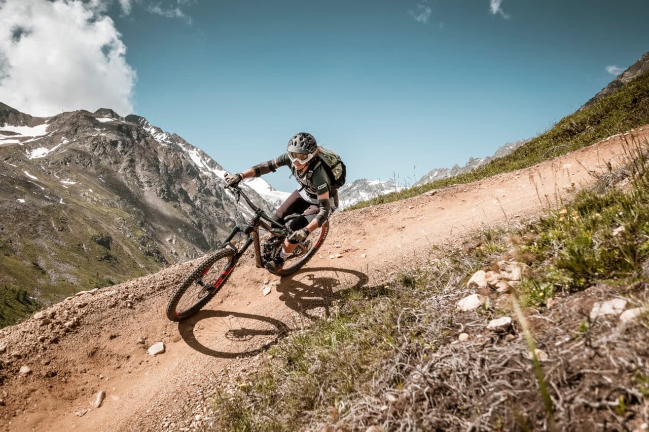 Ein Radfahrer mit Helm fährt mit einem Mountainbike einen Feldweg hinunter. Der Weg führt an einem steilen Hang entlang, mit felsigem Gelände und Bergen im Hintergrund unter einem klaren blauen Himmel.