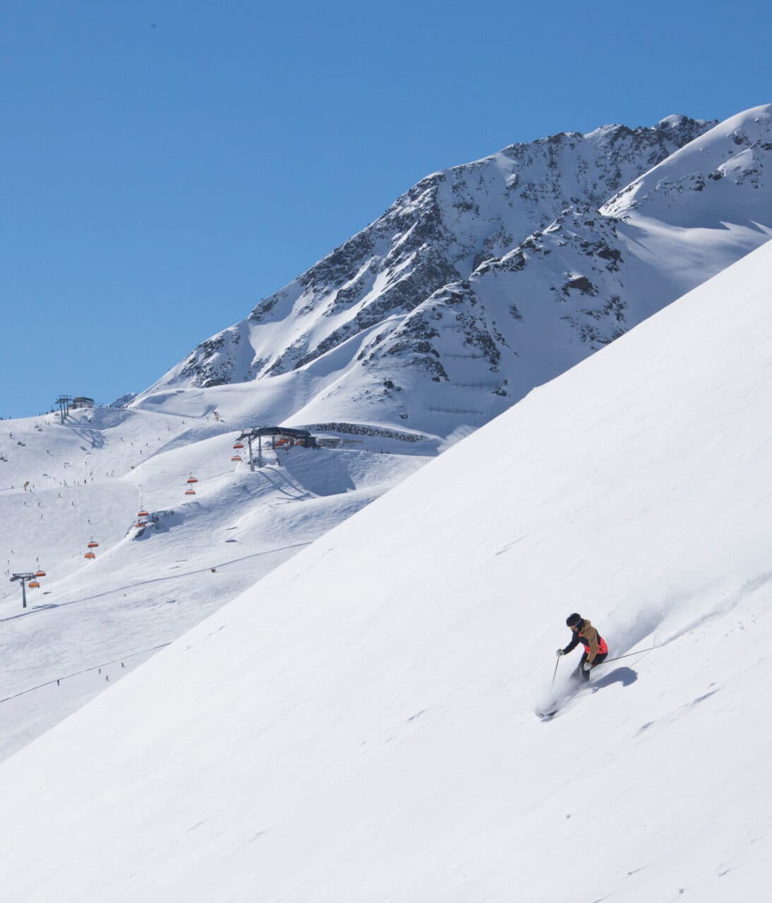 Person, die unter einem klaren blauen Himmel einen steilen, schneebedeckten Hang hinunterfährt, mit schneebedeckten Bergen und einem Skilift im Hintergrund.