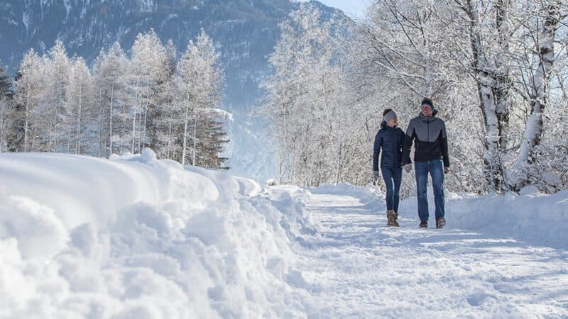 Ein Paar geht einen schneebedeckten, von Bäumen gesäumten Weg in einer bergigen Winterlandschaft entlang.