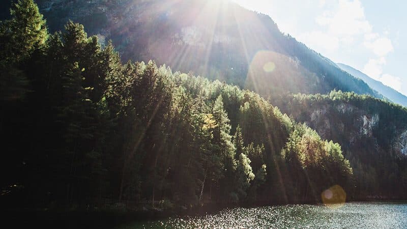 Sonnenlicht strömt über einen bewaldeten Berghang neben einem schimmernden See unter einem teilweise bewölkten Himmel.