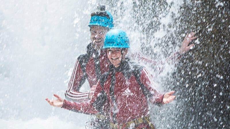 Zwei Menschen in roten Neoprenanzügen und blauen Helmen stehen lächelnd unter einem Wasserfall, um den sie vom Wasser umspült werden.