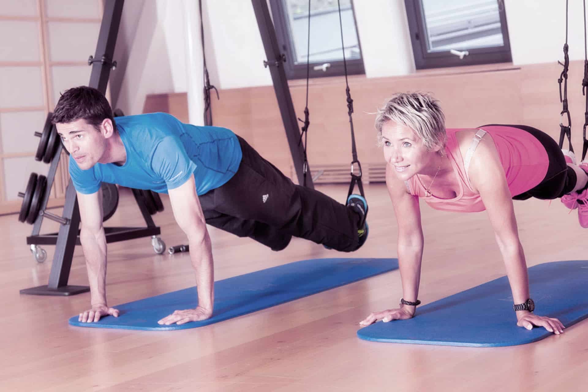 Zwei Personen machen Liegestütze auf blauen Matten in einem hell erleuchteten Raum, im Hintergrund sind Fenster und Trainingsgeräte zu sehen.