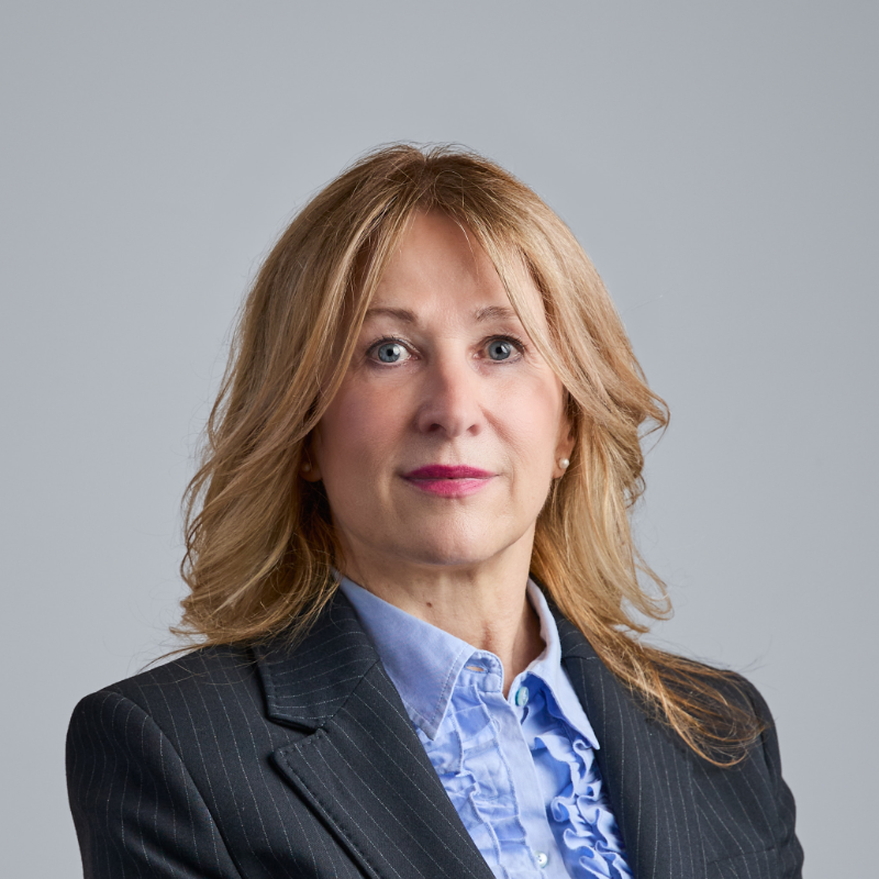 Eine Frau mit schulterlangen blonden Haaren, die einen dunklen, Nadelstreifenblazer über einer hellblauen Rüschenbluse trägt.