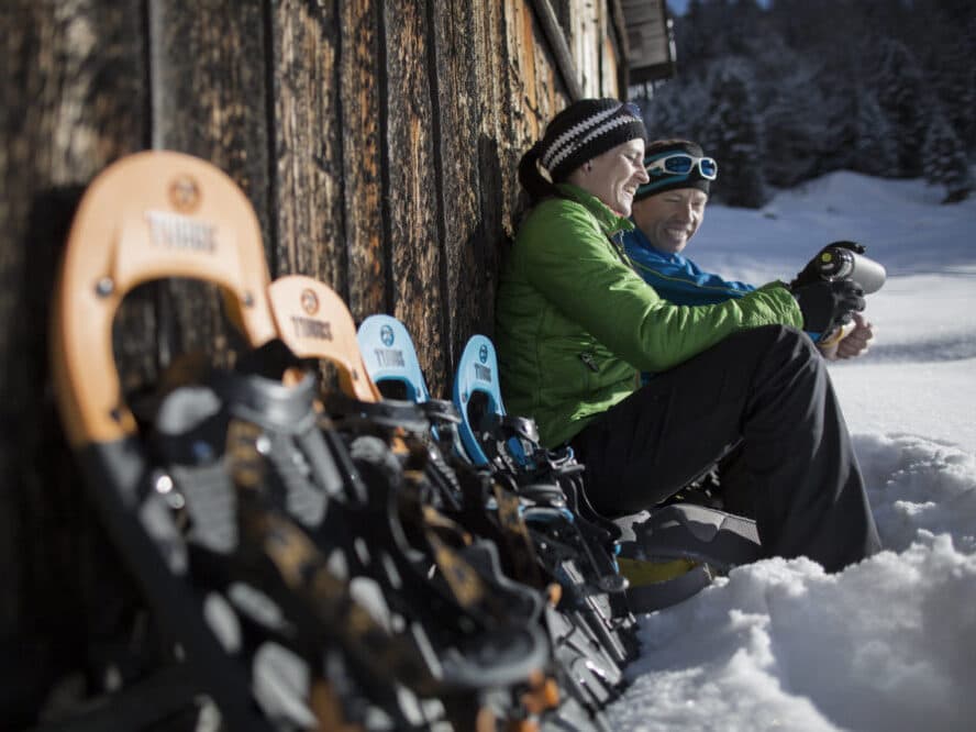 Two people sit against a wooden wall in the snow, wearing winter gear and adjusting snowshoes. Several snowshoes are lined up in the foreground.