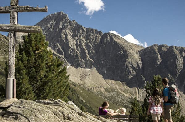 A family with their dog sits near a wooden cross on a rocky hill, taking in the rugged mountains beneath the clear summer sky.