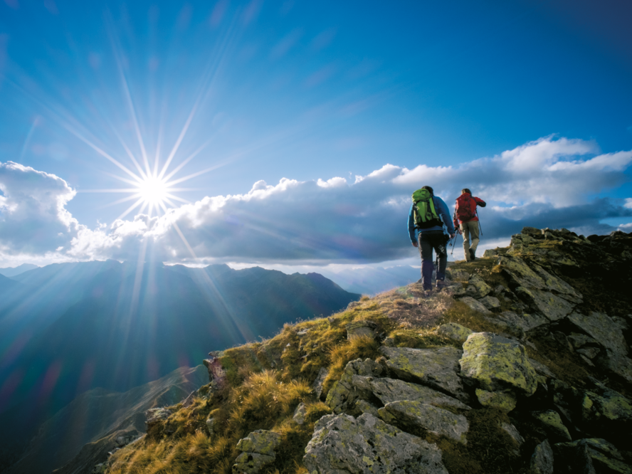 Two hikers with backpacks walk along a rocky mountain ridge under a bright sun and a partly cloudy sky.