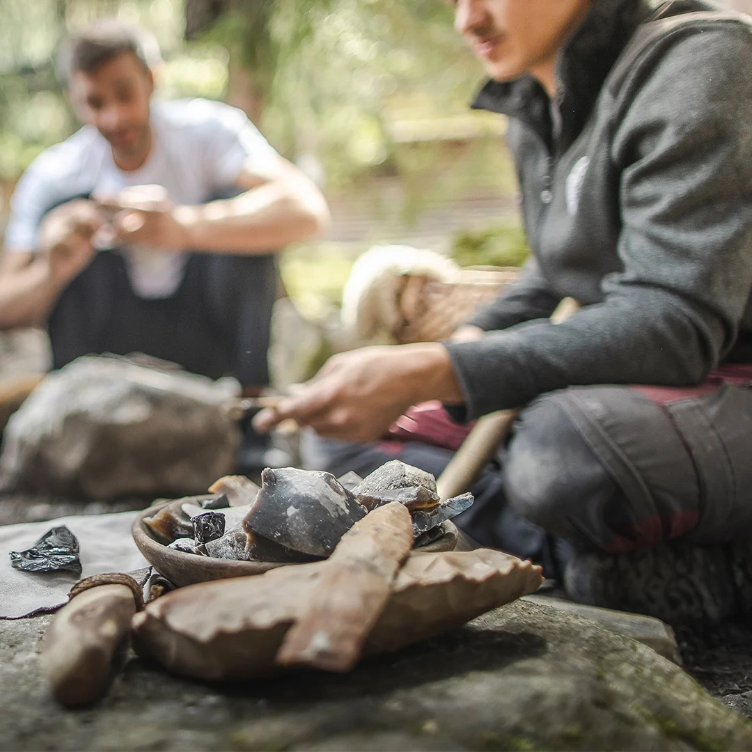 Zwei Personen sitzen im Freien und schlagen Feuersteine. Vor ihnen liegen verschiedene Steinwerkzeuge und Steine auf dem Boden ausgebreitet.