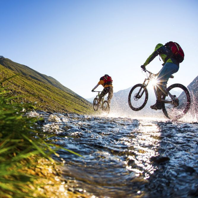 Zwei Radfahrer fahren an einem sonnigen Tag durch einen seichten Bach in einer bergigen Landschaft. Ein Radfahrer hebt sein Vorderrad an und spritzt Wasser auf.