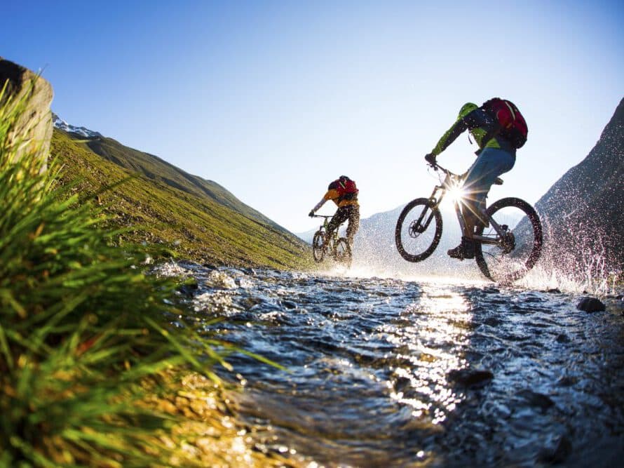 Two cyclists ride mountain bikes through a shallow stream, surrounded by green hills and a clear blue sky.