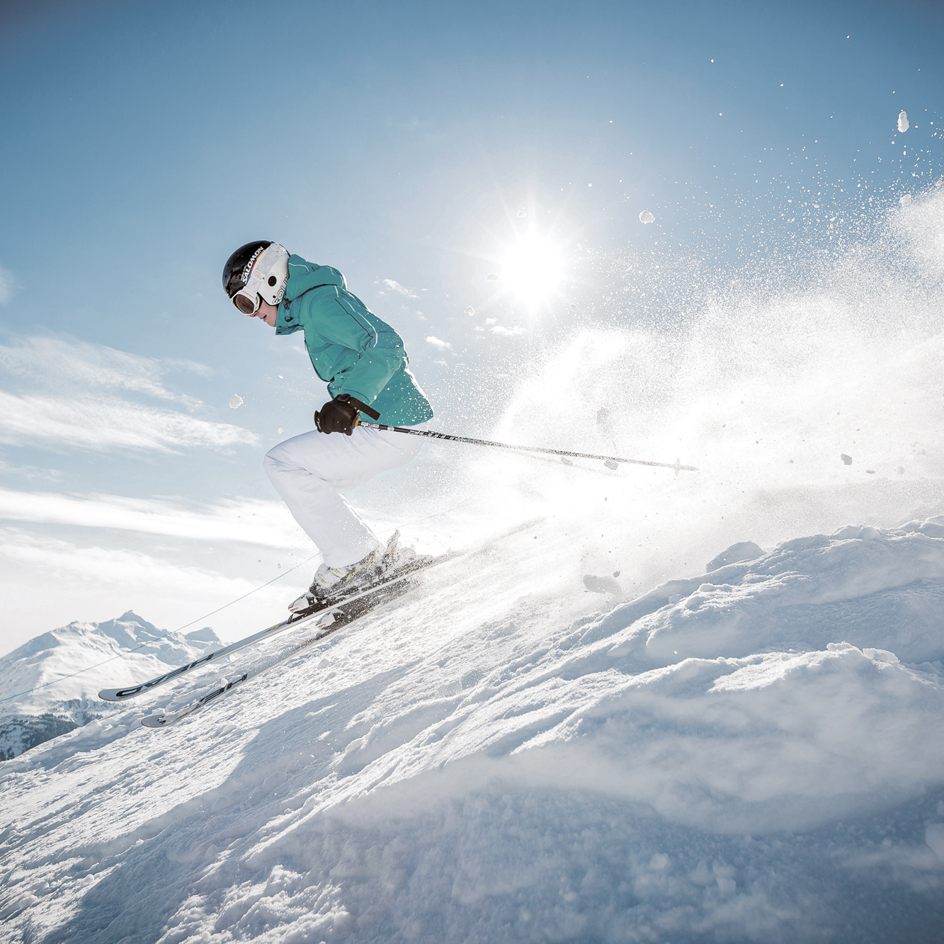 A skier in a green jacket and white pants descends a snowy slope under a clear sky with the sun shining brightly.