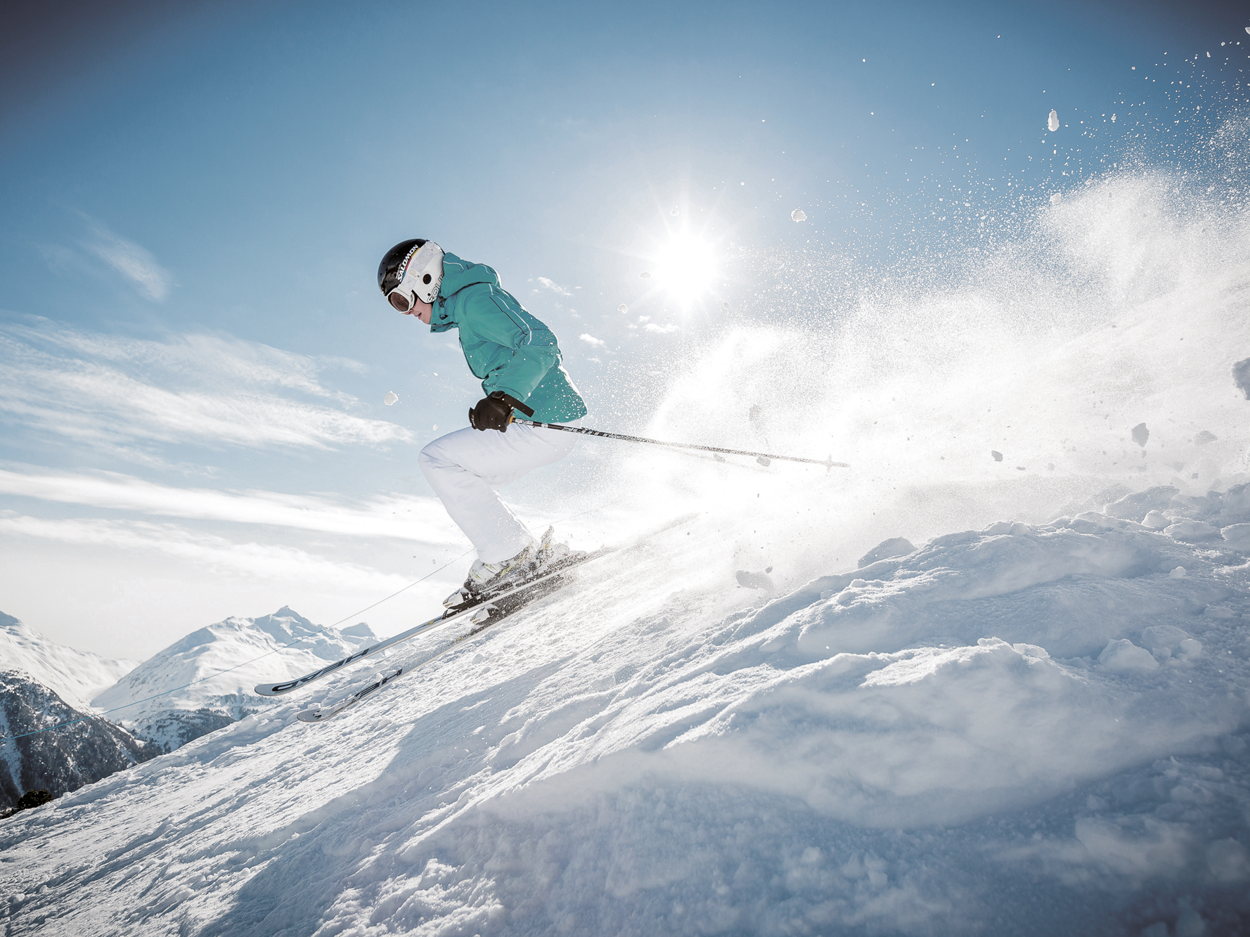 Ein Skifahrer in weiß-blaugrüner Ausrüstung fährt einen verschneiten Hang hinunter und erzeugt unter einem klaren blauen Himmel mit hellem Sonnenlicht eine Schneespritze.