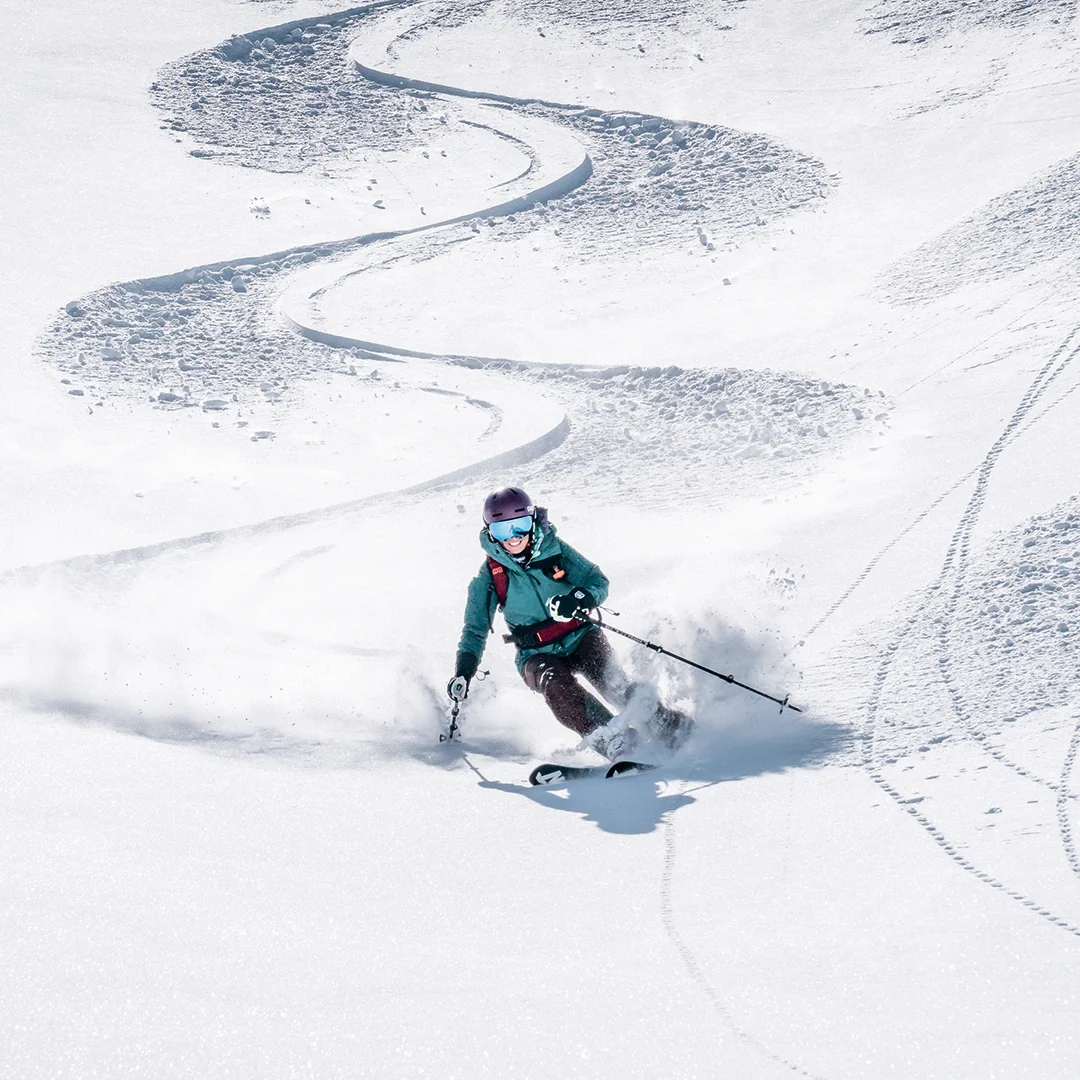 Eine Person in einer grünen Jacke und einem violetten Helm fährt auf Skiern den Neuschnee hinunter und zieht dabei eine Reihe S-förmiger Kurven.
