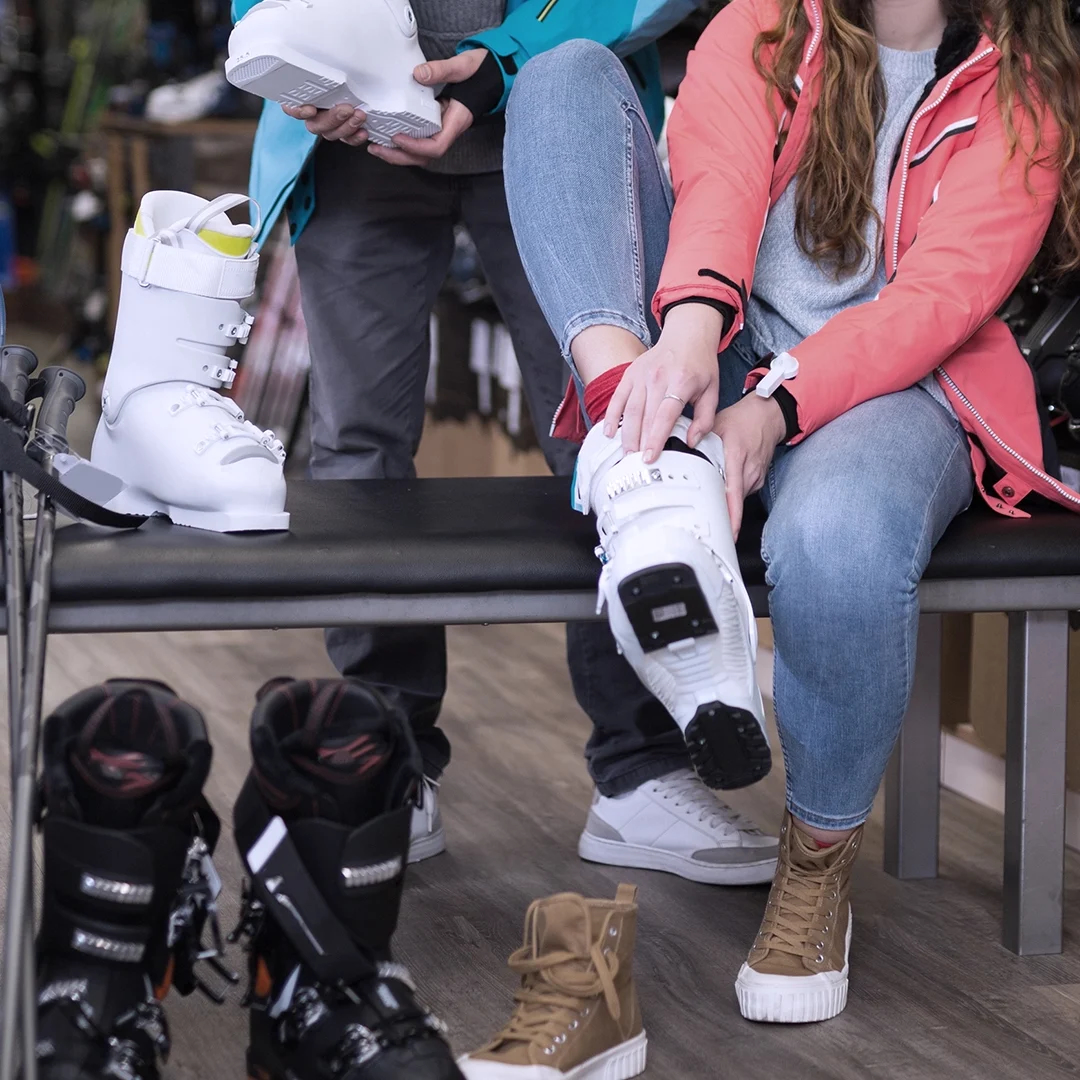 In einem geschäftigen Skiverleih sind zwei Freunde gerade dabei, Skischuhe anzuprobieren. Einer sitzt da und passt sorgfältig einen Schuh an, während der andere mit einem schicken Paar dasteht. Um sie herum liegen Freizeitschuhe und eine Auswahl an Skischuhen verstreut auf dem Boden und sorgen für eine lebhafte Atmosphäre voller Vorfreude und Aufregung.