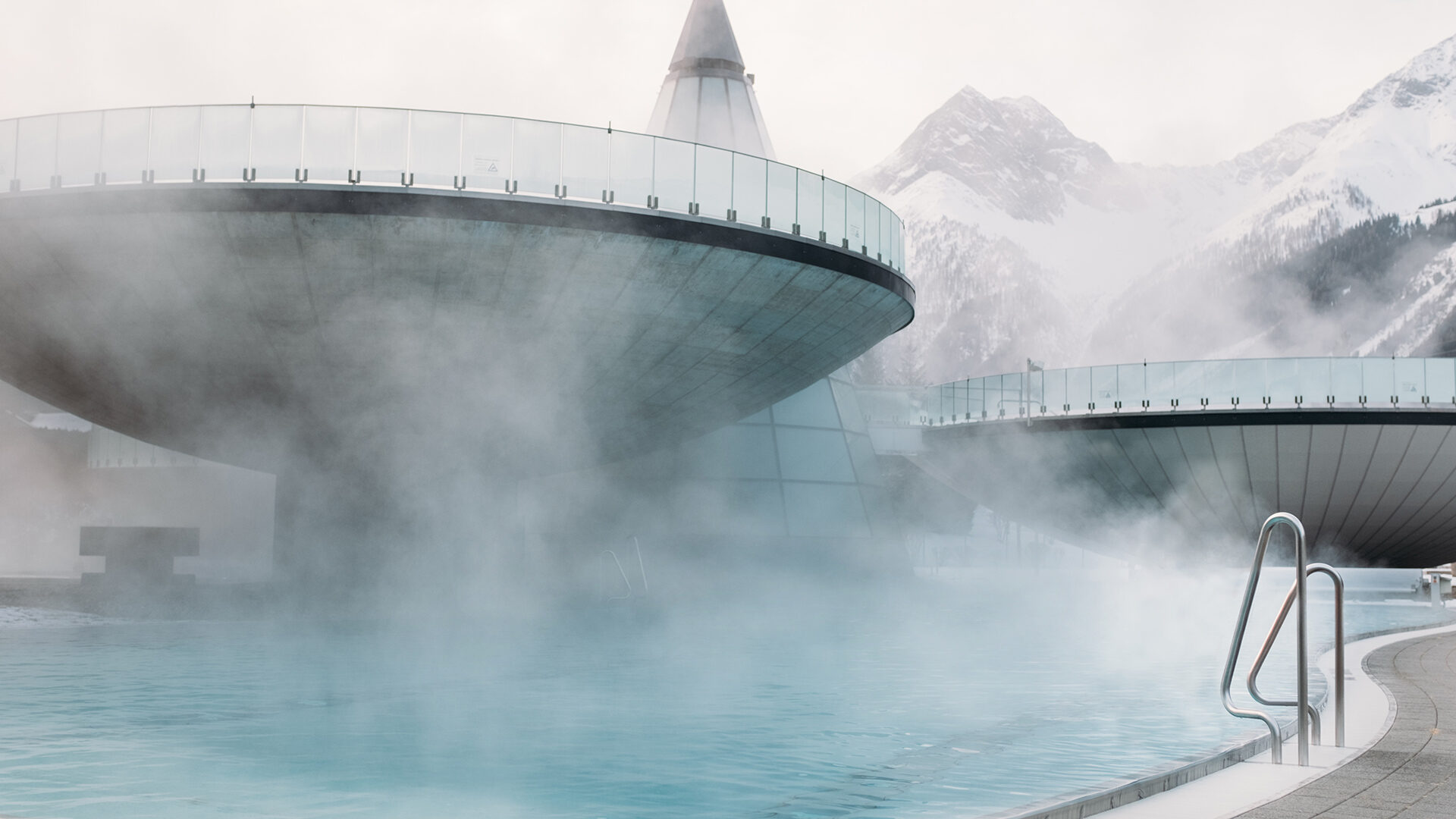 Der dampfumhüllte Außenpool liegt eingebettet zwischen den Strukturen der VAMED Vitality World vor der Kulisse schneebedeckter Berge. Ein schlanker Metallhandlauf erleichtert den Einstieg ins einladende Wasser.