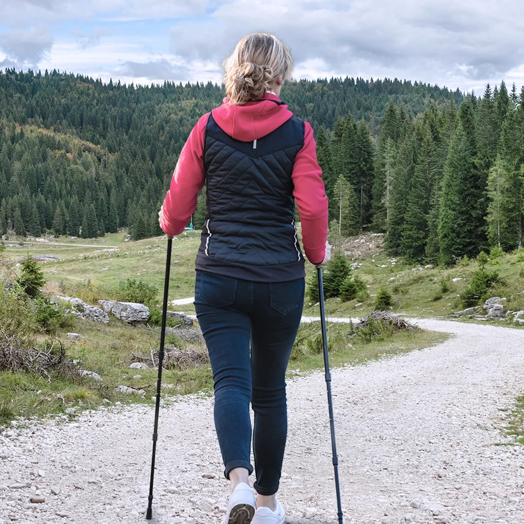 Eine Person mit Wanderstöcken geht unter bewölktem Himmel auf einem Schotterweg durch einen üppig grünen Wald.