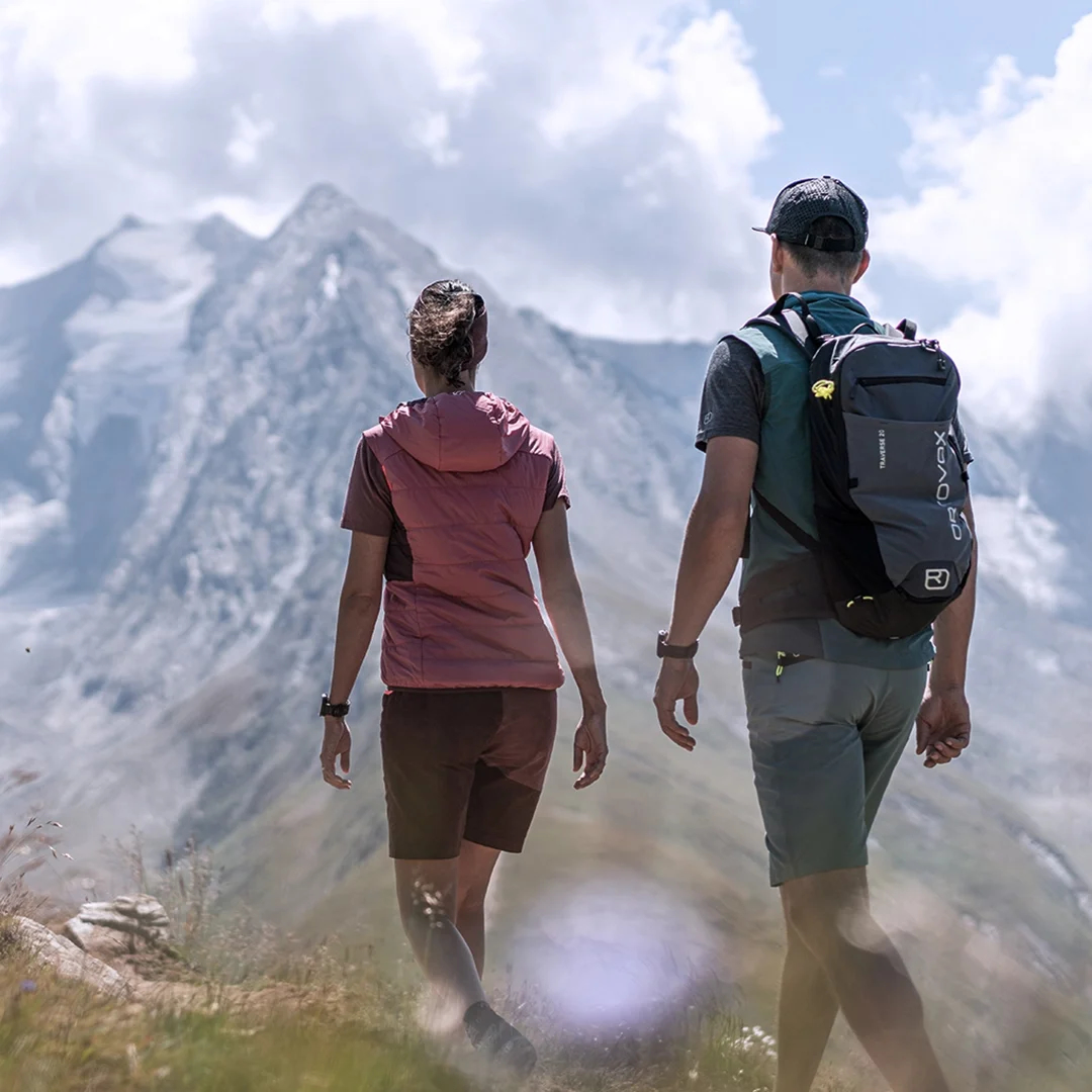 Zwei Wanderer gehen unter einem bewölkten Himmel auf einem Graspfad in Richtung schneebedeckter Berge.