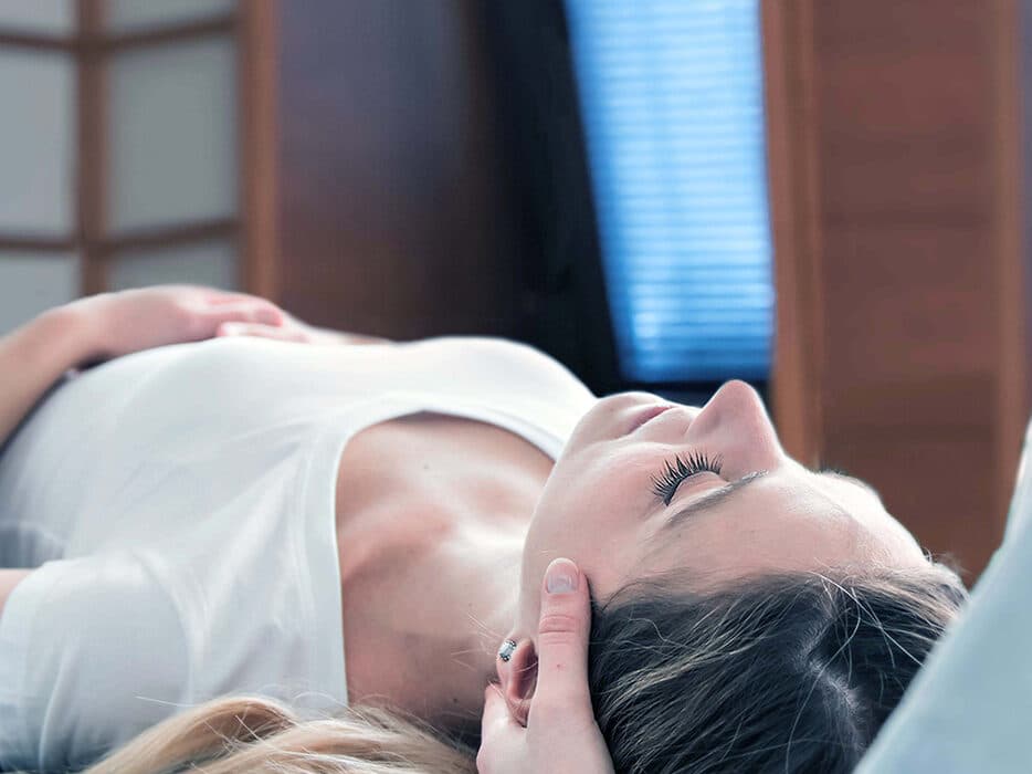 Eine Person liegt mit geschlossenen Augen und erhält eine Kopfmassage in einem Raum mit einem Holzschirm und einem Fenster.