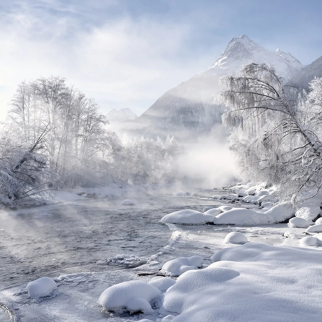 Eine verschneite Landschaft zeichnet sich durch einen gewundenen Fluss aus, der von schneebedeckten Bäumen gesäumt ist. Im Hintergrund erhebt sich ein Berggipfel unter einem klaren Himmel, über dem Nebel liegt.