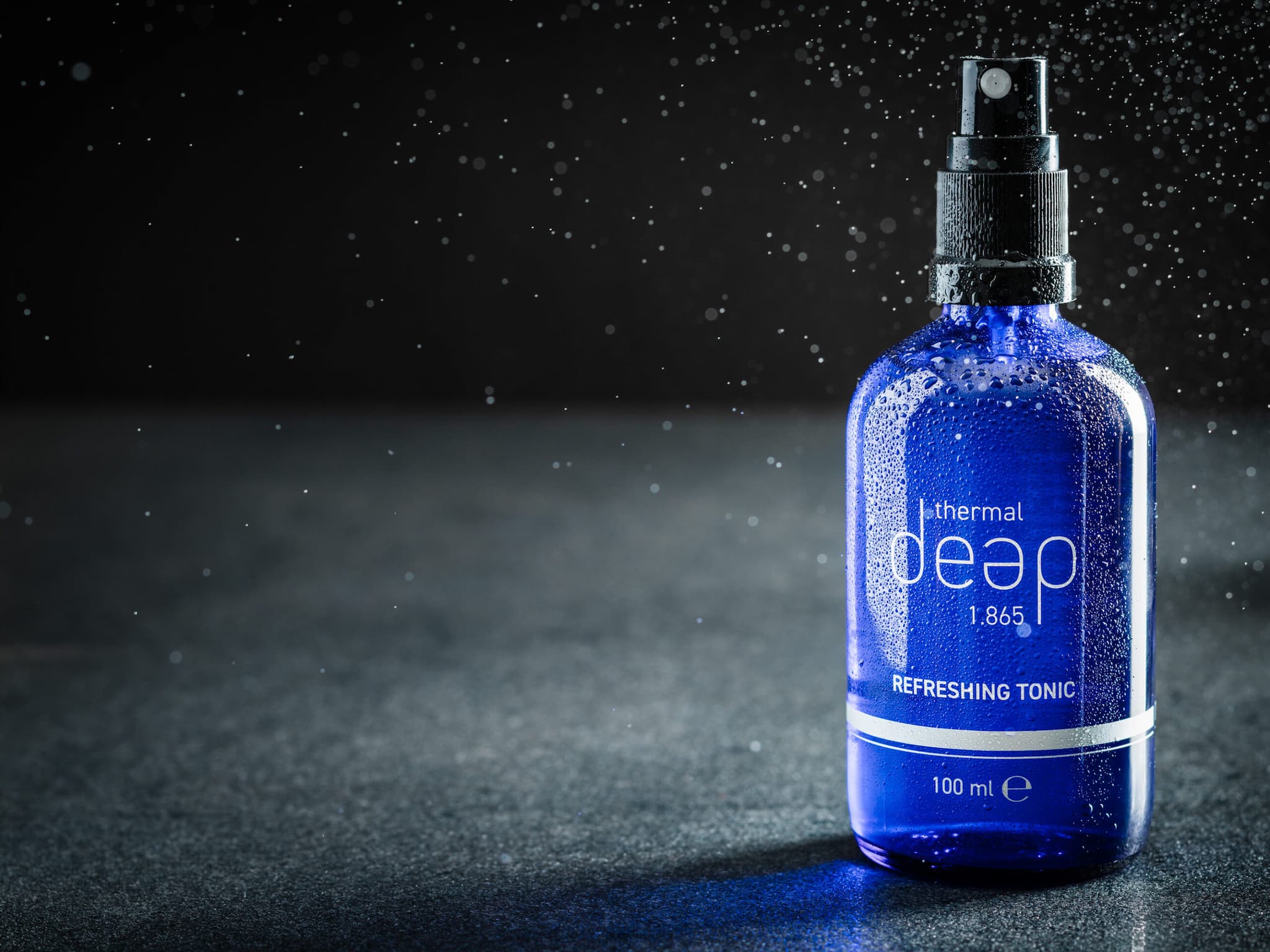 Eine blaue Flasche „Deep 1.865 Refreshing Tonic“ mit Sprühdüse steht auf einer dunklen Oberfläche und schafft inmitten schwebender Wassertropfen eine Atmosphäre der Verjüngung und ruft das erfrischende Gefühl thermischer Tiefen hervor.