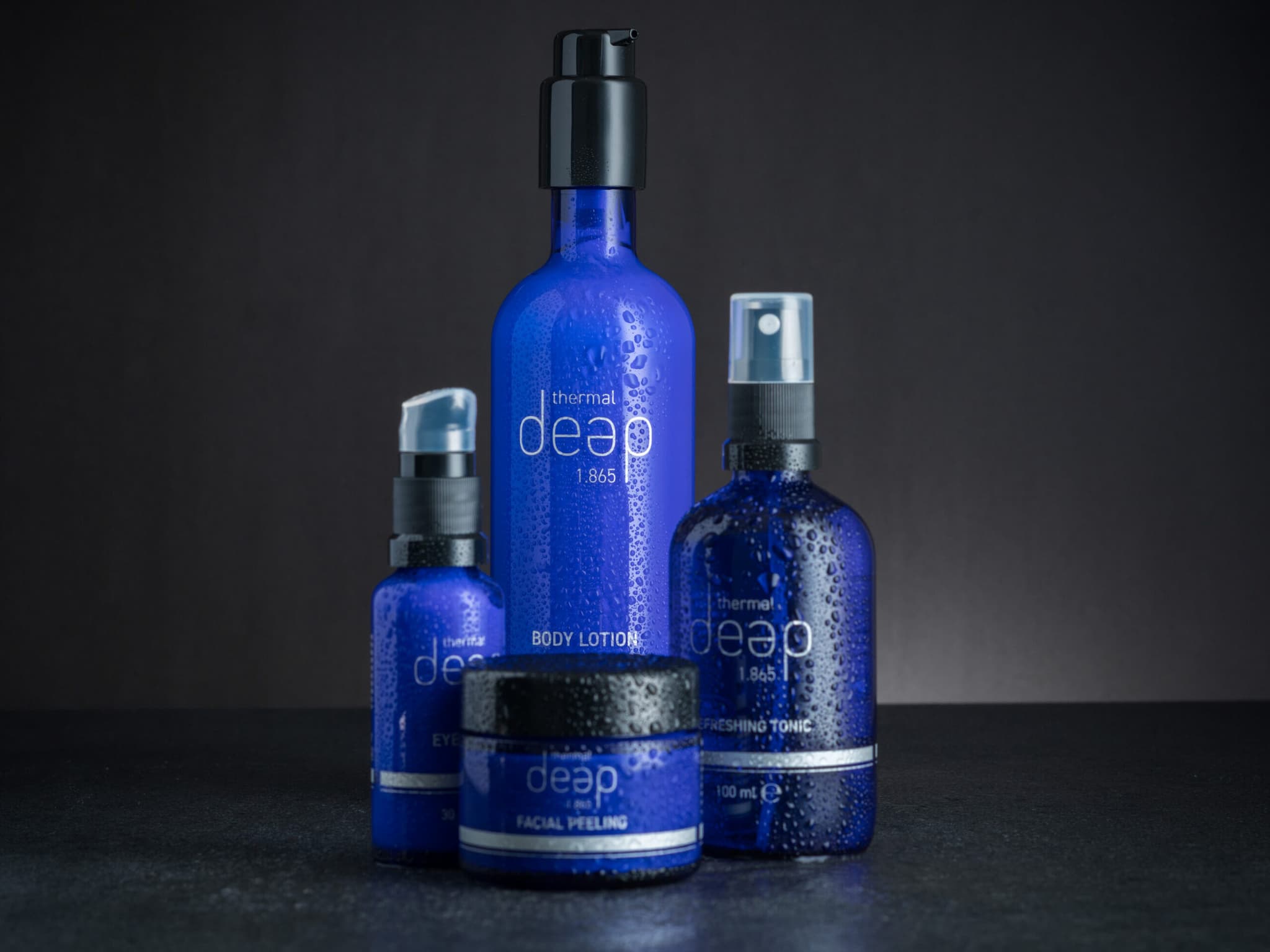 Eine Kollektion blauer Hautpflegeprodukte mit thermischen Eigenschaften, darunter Körperlotion, erfrischendes Tonic, Augenserum und Gesichtspeeling. Jedes Produkt ist auf einer dunklen Oberfläche präsentiert, auf deren Flaschen glitzernde Wassertropfen für einen frischen Look sorgen.