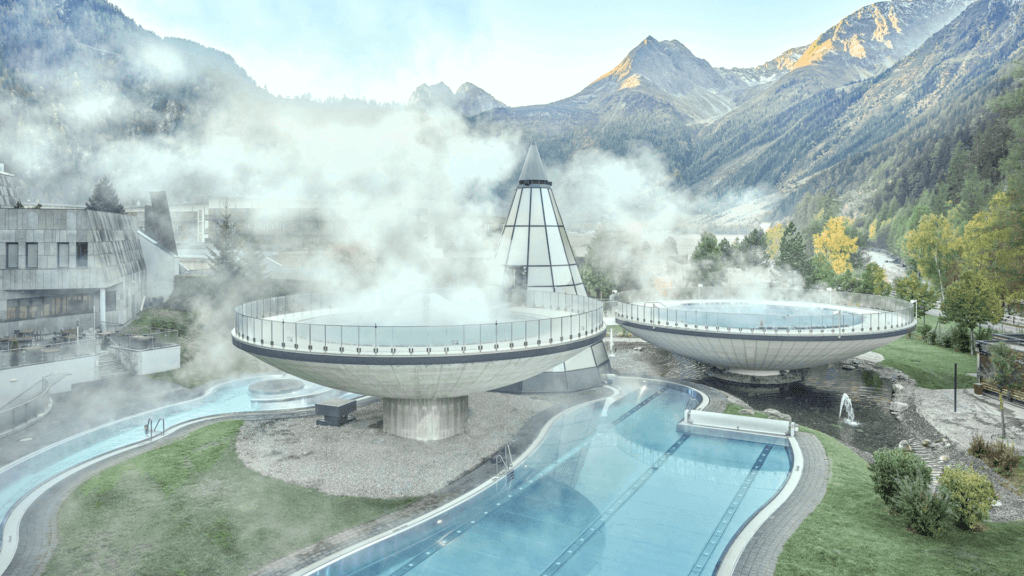 Eingebettet in eine Berglandschaft laden die futuristischen Kurgebäude zum Thermenurlaub ein. Elegant steigt Dampf aus ihren schlanken Strukturen auf und vermischt sich mit dem Nebel, der das Außenbecken umhüllt, und sorgt für eine ätherische Auszeit der Entspannung.