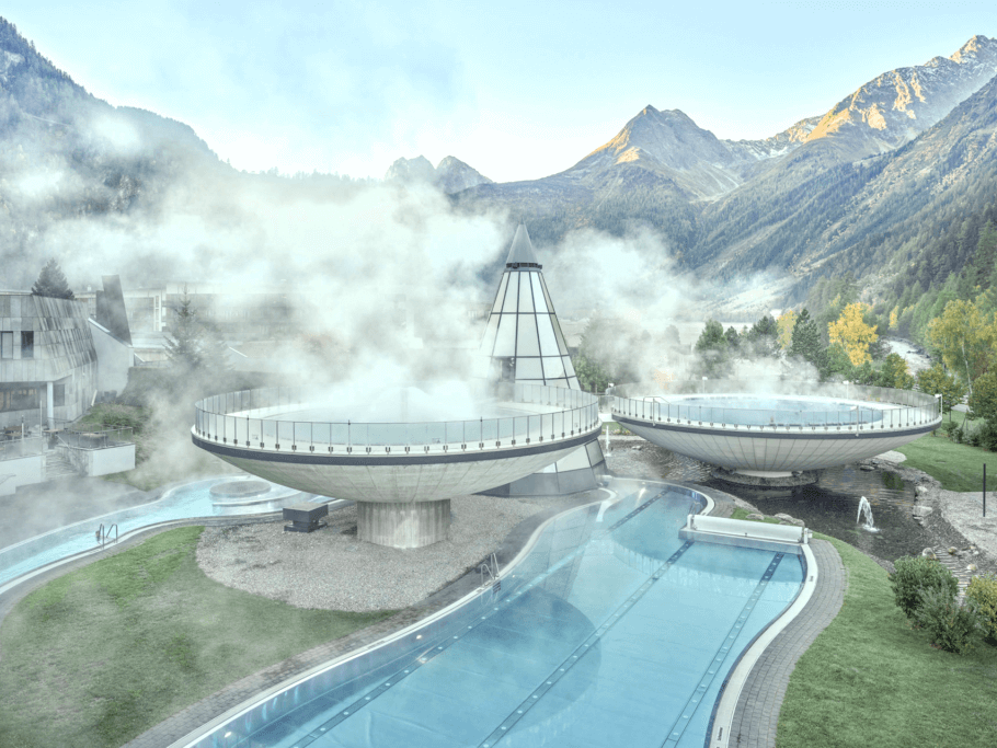 Futuristisches Spa mit dampfenden, runden Pools, umgeben von Bergen und Bäumen. Im Vordergrund befindet sich ein klarer Pool, um den herum Nebel aufsteigt.
